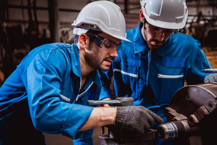 Zwei Männer in blauen Arbeitsanzügen und weißen Schutzhelmen, die an einer Metallmaschine arbeiten. Beide tragen Schutzbrillen und Handschuhe. Sie befinden sich in einer industriellen Werkstattumgebung.  